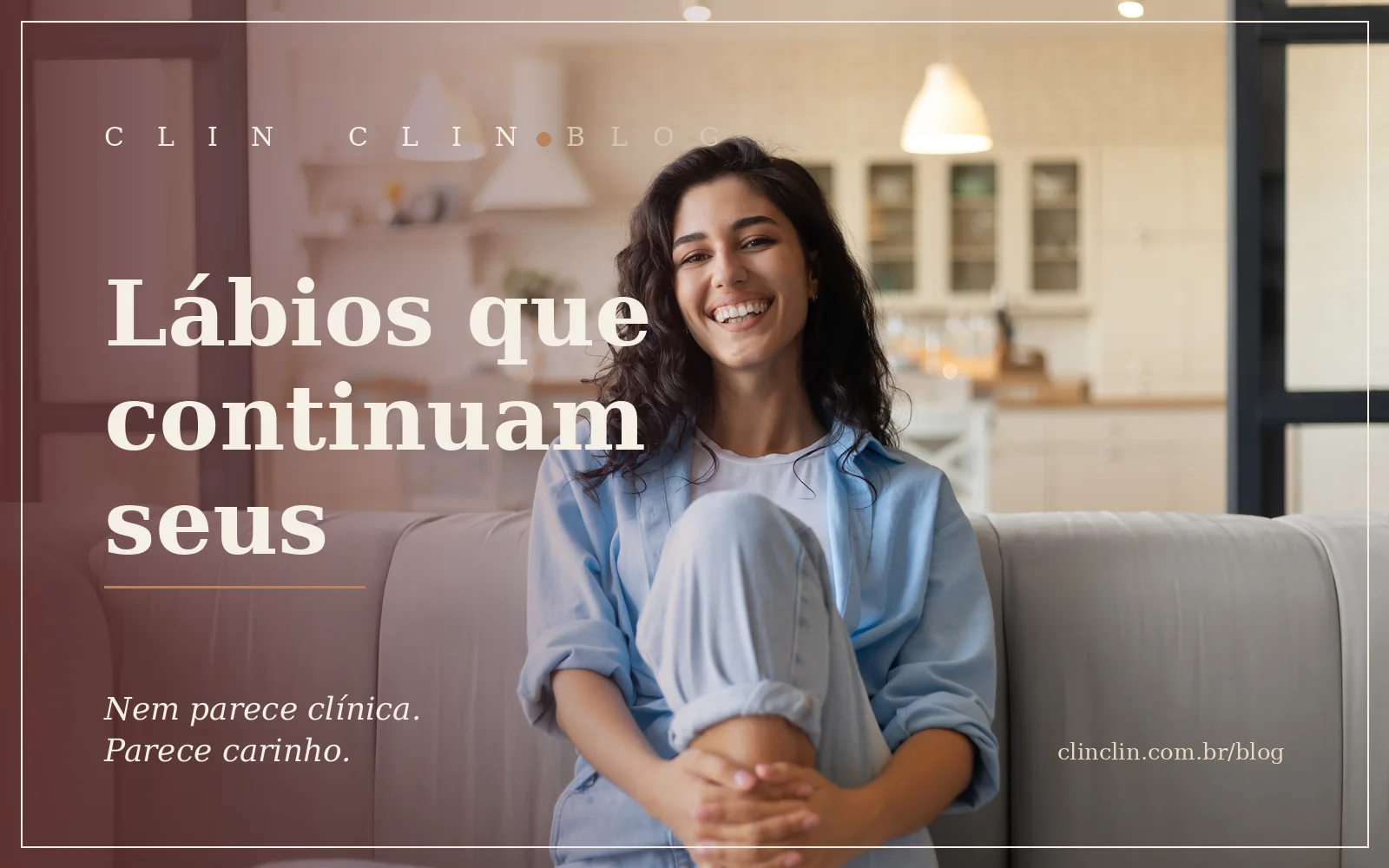 Mulher jovem morena de cabelos ondulados, sorriso espontâneo, sentada no sofá de casa, de camisa azul-clara, sob luz natural suave